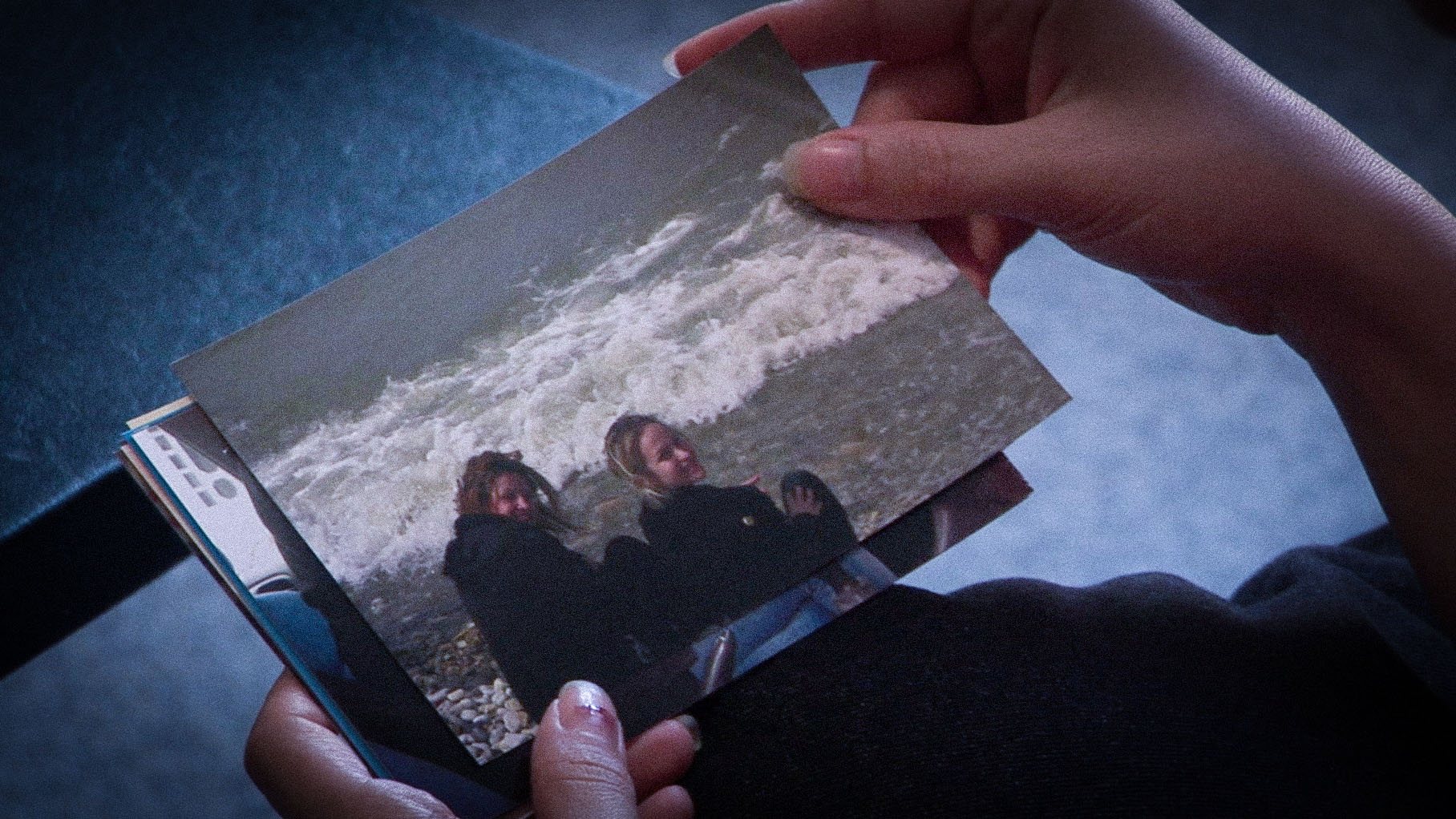 Hands hold a printed photograph of two young women sitting on a beach in front of breaking waves.