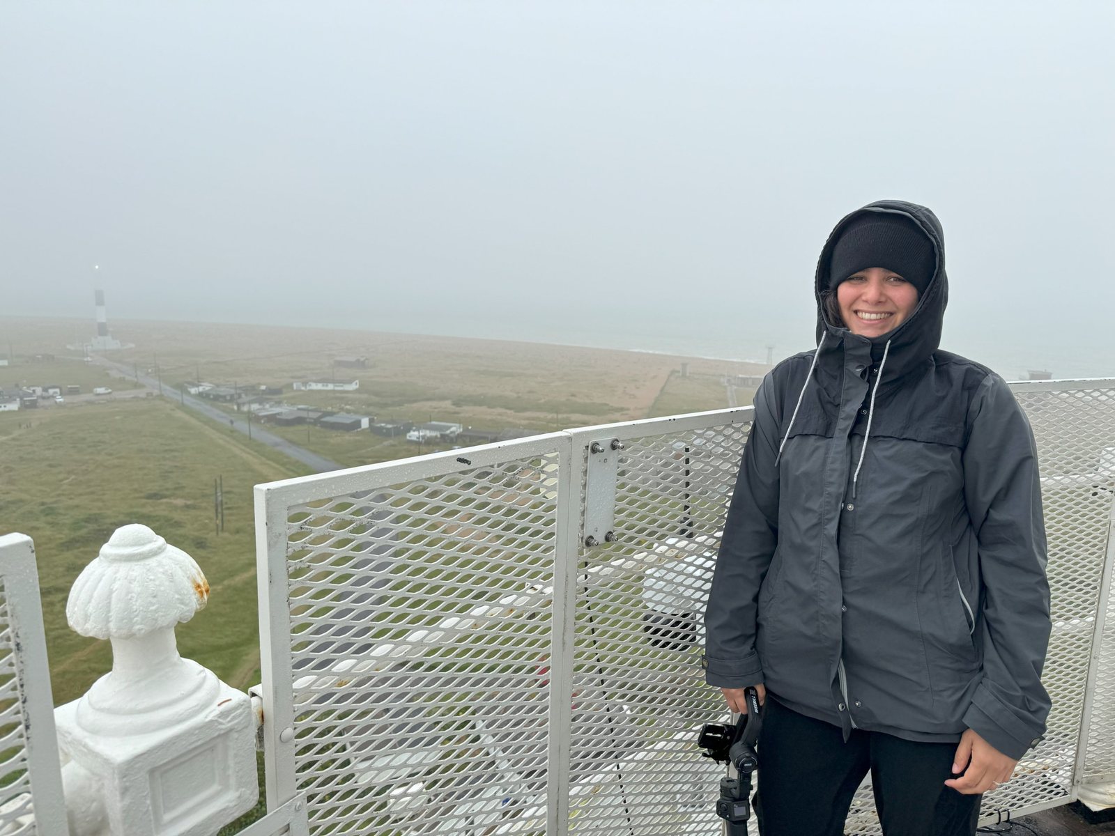 Mariana Duarte with a camera gimbal on the Old Lighthouse gallery at Dungeness, with the new lighthouse and the shingle beach behind