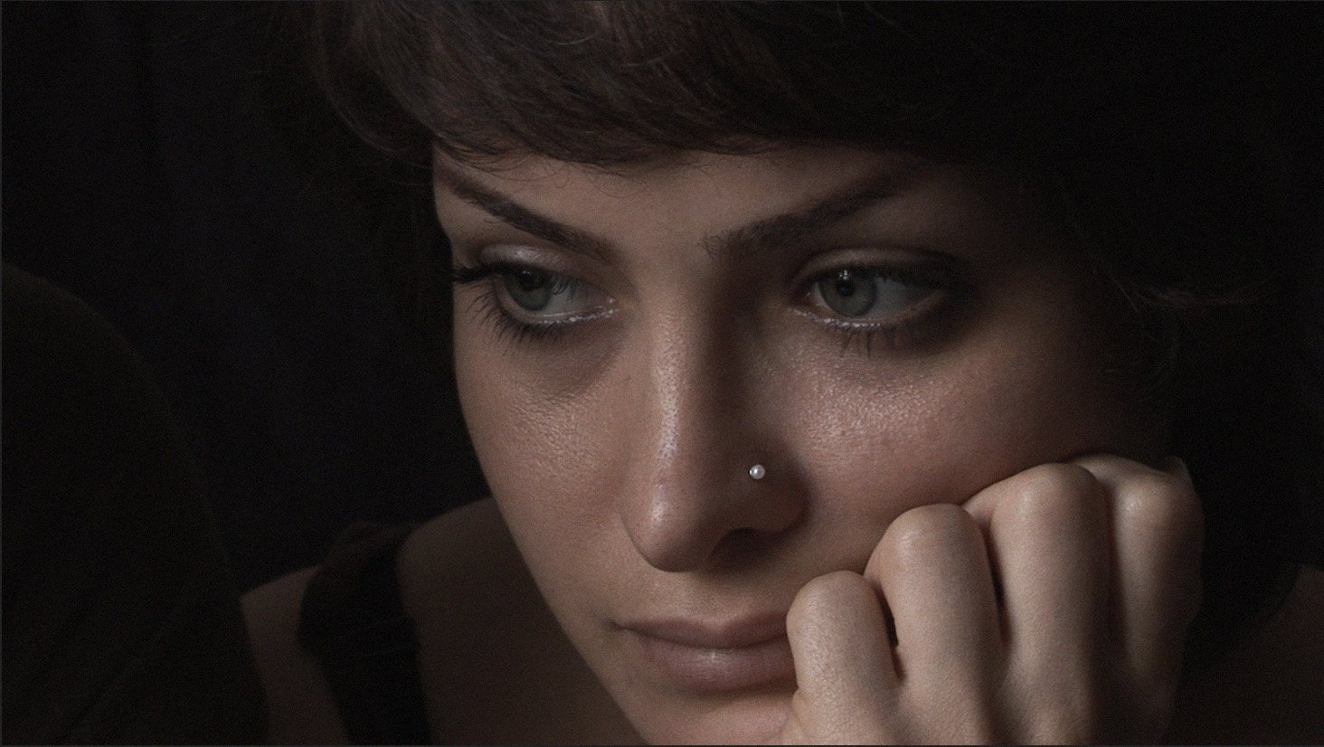 Close-up of a young woman resting her chin on her hand, looking down, her face lit softly against a dark background.