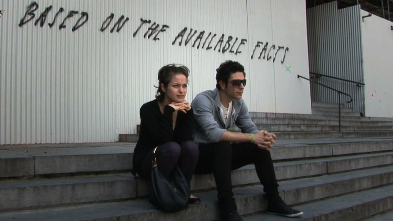 The couple sit on concrete steps below graffiti that reads 'Based on the available facts'.