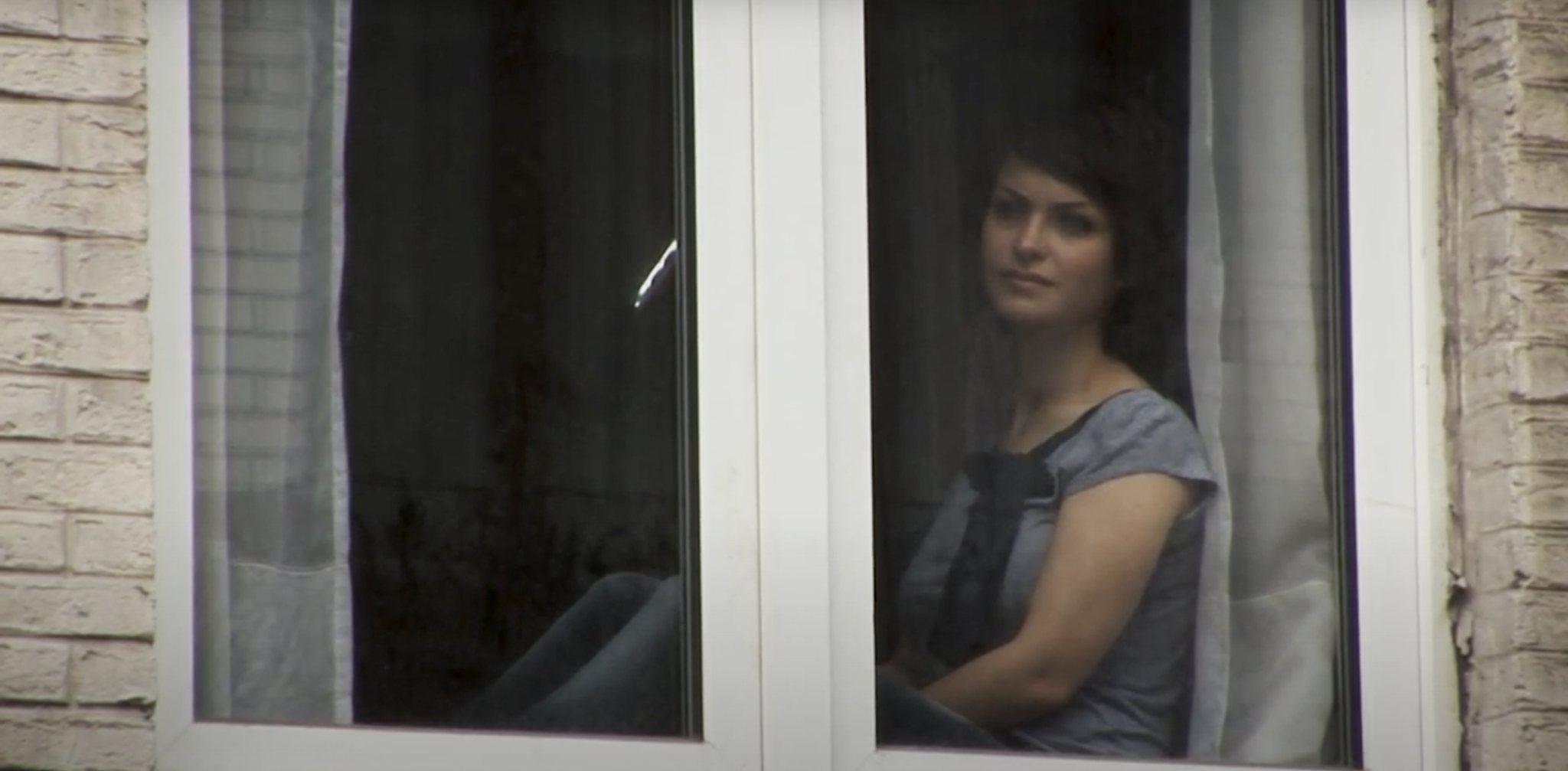 A young woman sits behind a window, looking out at the street.